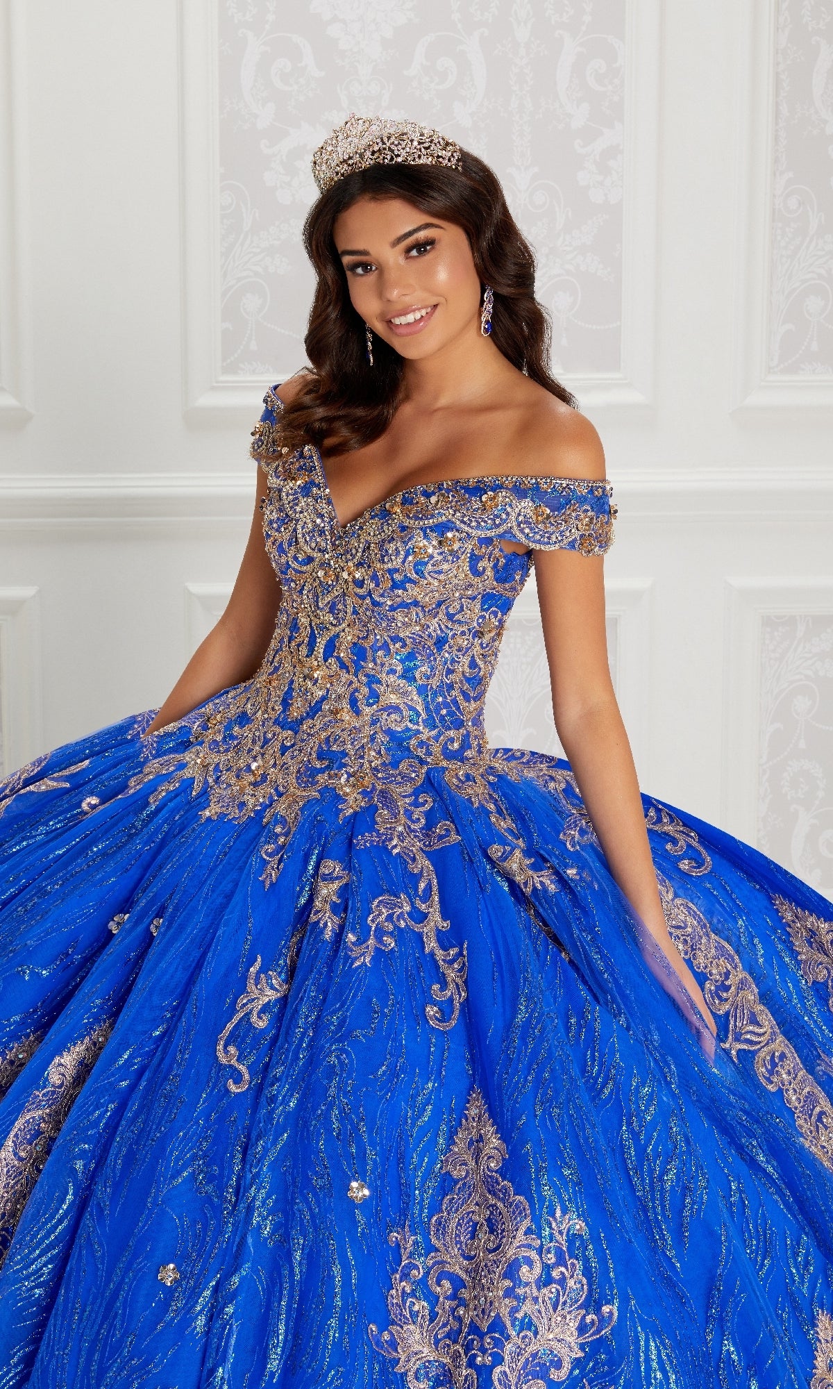 A young woman in the Quinceanera Dress PR22148 By Princesa, featuring sparkling blue and gold beaded accents with a jeweled tiara, poses against an ornate white background.